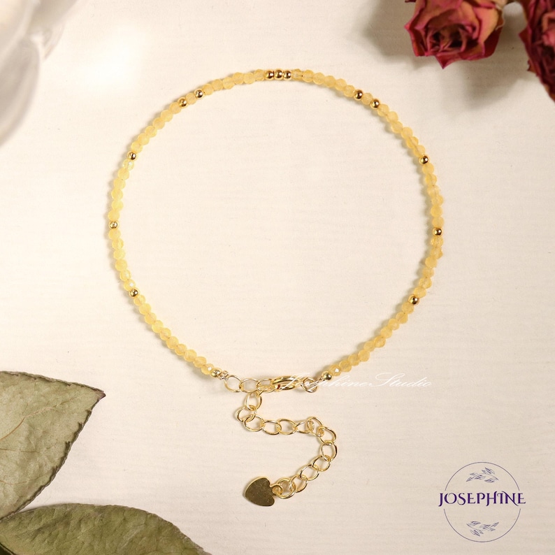 May include: A delicate bracelet featuring small, faceted pale yellow beads interspersed with tiny gold-toned metal spheres. The piece includes a gold-toned chain extender with a small heart-shaped charm at the end and a lobster clasp closure.