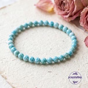 May include: A light blue beaded bracelet with a pattern of white spots. The bracelet is made of round beads and is displayed on a textured, off-white surface. The word "JOSEPHINE" is in a circle at the bottom right.