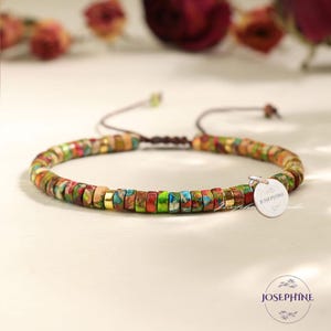 Könnte beinhalten: Ein farbenfrohes Perlenarmband mit einem silbernen Anhänger, auf dem "Josephine" steht. Das Armband besteht aus kleinen, runden Perlen in verschiedenen Grüntönen, Blau, Rot und Braun. Die Perlen sind auf einer braunen Schnur aufgefädelt und am Ende mit einem Knoten befestigt.