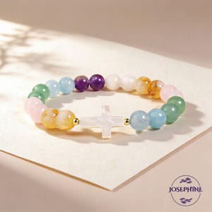 May include: A beaded bracelet featuring a white cross pendant and a variety of colorful gemstone beads. The beads include pink, green, yellow, purple, and blue hues. The bracelet is displayed on a light-colored surface.