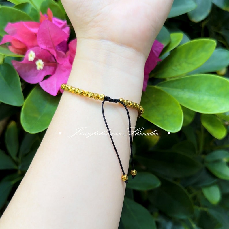 May include: A gold bracelet with a black cord and gold beads. The bracelet is tied with a knot and has a tassel.