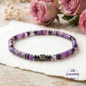 Könnte beinhalten: Ein Armband mit Amethyst- und dunkelgrauen Hämatitperlen. Das Armband hat eine zentrale geometrische Perle. Es wird auf einer weißen Oberfläche mit floralem Hintergrund präsentiert. Das Armband ist ein Schmuckstück.