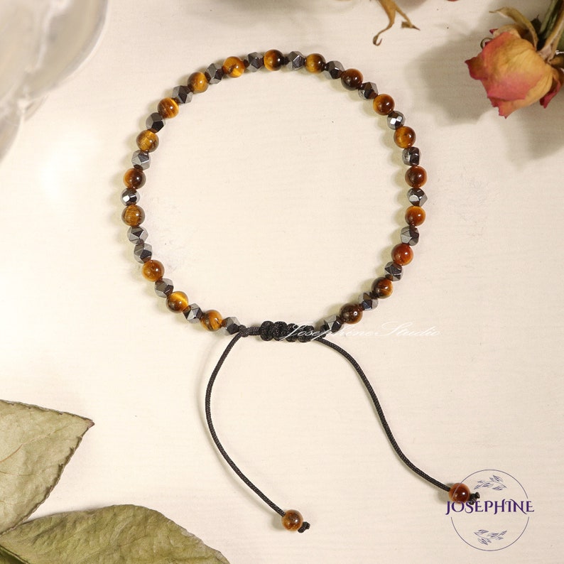 May include: A brown and black beaded bracelet with a black cord and adjustable clasp. The bracelet is made with tiger's eye and hematite beads.