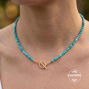 May include: A beaded necklace featuring small, round, turquoise-colored stones with natural veining, accented by a gold-toned metal toggle clasp at the center. The necklace rests against the neck, showcasing a consistent pattern of beads.