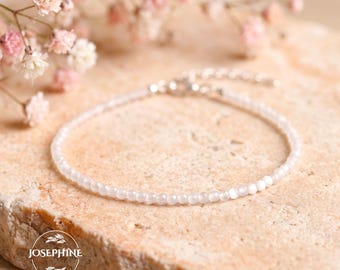 May include: A delicate bracelet composed of small, translucent white beads with a subtle sheen. The bracelet features a silver-toned metal clasp and an adjustable chain extension. A circular logo with the text JOSEPHINE is visible.