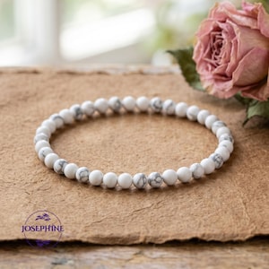 May include: White and gray beaded bracelet. The bracelet is made of small, round beads with a marbled gray pattern. The bracelet is displayed on a brown surface, with a pink rose in the background. The brand name "Josephine" is visible.