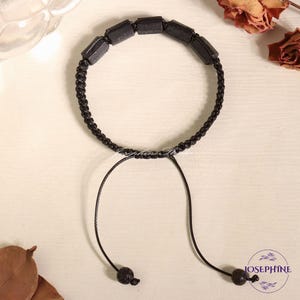 May include: Black beaded bracelet with a macrame cord and adjustable closure. The bracelet features rectangular black beads and a woven cord. The adjustable closure has two black, round beads. The bracelet is on a light-colored surface.