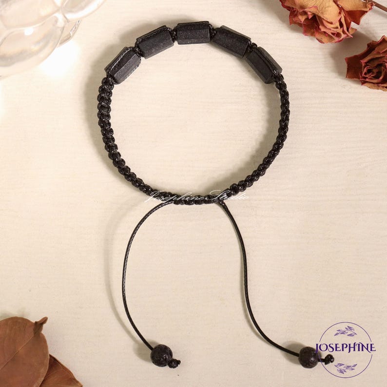 May include: Black beaded bracelet with a macrame cord and adjustable closure. The bracelet features rectangular black beads and a woven cord. The adjustable closure has two black, round beads. The bracelet is on a light-colored surface.