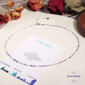 May include: A delicate necklace made with small, iridescent purple and clear beads. The necklace is on a white card with the text "Fluorite is renowned for its cleansing energy, focus-enhancing abilities, and soothing effects on the mind and emotions." and the text "Fluorite" in a larger font. The card also has the text "Cleansing", "Focus", and "Calming" with small crystal images.