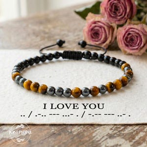 May include: A beaded bracelet with black, gray, and amber-colored beads. The bracelet is on a white surface with the text "I LOVE YOU" and Morse code. The word "JOSEPHINE" is in a circle at the bottom.