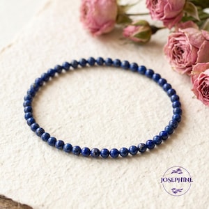 May include: A delicate bracelet composed of small, round, deep blue beads. The beads are strung together to form a complete circle. The bracelet is displayed on a textured, off-white surface, with dried pink roses in the background. The brand logo "JOSEPHINE" is visible.