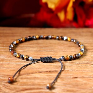 May include: A brown and gold beaded bracelet with a black cord and adjustable clasp. The bracelet is made of tiger's eye and hematite beads.