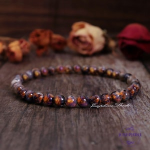 Puede incluir: Un brazalete hecho de pequeñas cuentas redondas multicolores. Las cuentas son una mezcla de marrón, naranja y morado con un patrón moteado. El brazalete está sobre una superficie de madera.