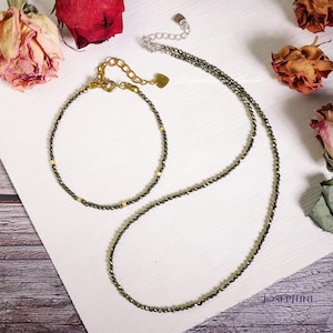 Puede incluir: Un conjunto de dos collares hechos de pequeñas cuentas de color gris oscuro con detalles dorados. El collar más largo tiene un dije en forma de corazón. El collar más corto es una pulsera.