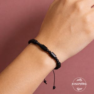 May include: Black beaded bracelet with rectangular and round beads. The bracelet is on a wrist against a pink background. The bracelet is adjustable with a black string.