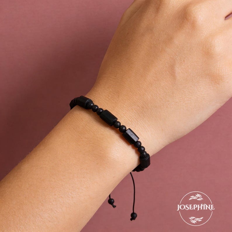May include: Black beaded bracelet with rectangular and round beads. The bracelet is on a wrist against a pink background. The bracelet is adjustable with a black string.