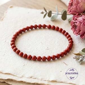 May include: A red beaded bracelet with small, round beads. The bracelet is displayed on a stack of textured, off-white paper. Dried pink roses and greenery are in the background. The bracelet is a jewelry item.