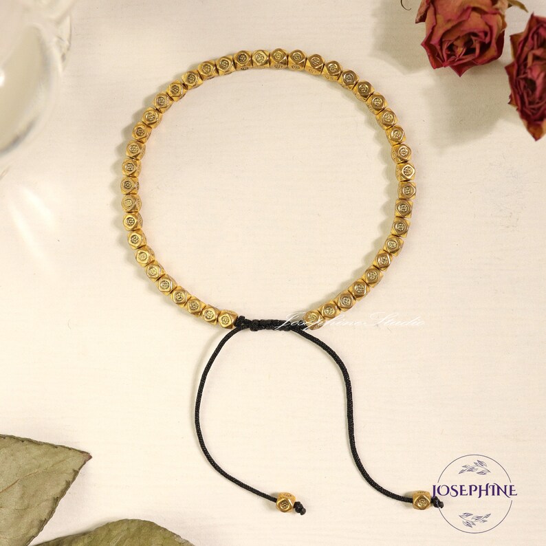 May include: A gold bracelet with a black cord and a sliding knot closure. The bracelet is made of small, round beads with a raised design. The bracelet is on a white surface.