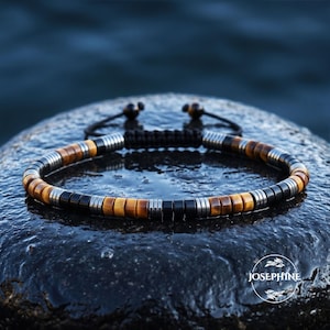 Puede incluir: Una pulsera de cuentas con cuentas cilíndricas alternas de ojo de tigre marrón, negro mate y metal plateado. La pulsera incluye un cierre de cordón negro con dos pequeñas cuentas de ojo de tigre en los extremos y el texto JOSEPHINE en blanco.