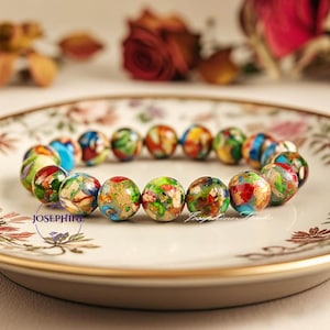 May include: A colorful bracelet made of round beads with a variety of patterns and colors, including blue, green, red, and gold. The bracelet is on a white plate with a floral design.