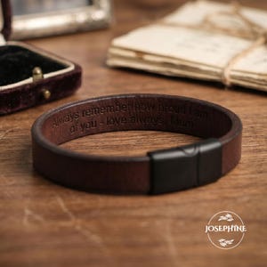 May include: A dark brown leather bracelet featuring a matte black metal clasp. The interior of the leather band is engraved with the text: Always remember how proud I am of you - love always, Mum. The bracelet has a smooth, simple design.