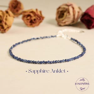 Puede incluir: Una tobillera delicada con pequeñas cuentas de zafiro azul oscuro facetadas dispuestas en una sola hebra. La pieza incluye un cierre de metal plateado y una cadena extensora ajustable. El texto Sapphire Anklet aparece impreso debajo.