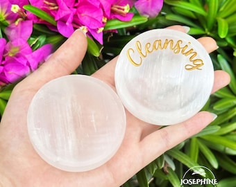May include: Two round, white, translucent selenite crystal bowls. One bowl is plain, while the other features the word Cleansing written in gold cursive script on its surface. The bowls have a smooth, polished finish and a circular shape.