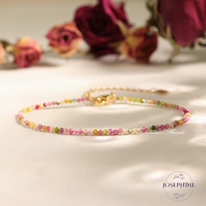 May include: A delicate bracelet made of small, colorful gemstones. The bracelet is made of pink, yellow, green, and blue gemstones. The bracelet is on a white surface.