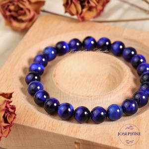 May include: A beaded bracelet featuring polished, deep blue tiger eye gemstones. The bracelet is displayed on a light wooden tray. The gemstones are approximately 0.4 inches in diameter. The bracelet is a piece of jewelry.