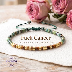 May include: A beaded bracelet featuring green and brown cylindrical beads with gold-tone spacers, tied with a dark green cord. The text "Fuck Cancer" is printed above a row of braille dots. A circular logo with the text "JOSEPHINE" is visible.