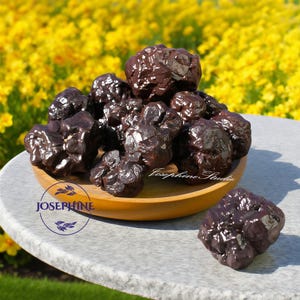 May include: A collection of dark brown, clustered mineral specimens displayed in a round wooden bowl. The minerals have a shiny, crystalline appearance. The bowl sits on a gray stone surface, with a blurred background of yellow flowers.