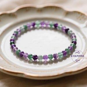 Könnte beinhalten: Ein Armband aus runden Perlen in Lila, Grün und transparenten Farben. Das Armband liegt auf einem weißen Teller mit gewelltem Rand. Das Wort "JOSEPHINE" ist auf dem Teller aufgedruckt.