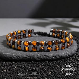 Könnte beinhalten: Armband mit Tigerauge- und grauen Hämatit-Perlen. Das Armband ist auf einem schwarzen Kordelband aufgefädelt und liegt auf einem dunkelgrauen Stein. Das Logo von Josephine Studio ist sichtbar.