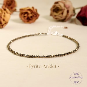 Puede incluir: Una delicada pulsera de tobillo de pirita con pequeñas cuentas facetadas en tonos dorados y bronce. La pulsera se muestra sobre una superficie clara, con flores secas en el fondo. El texto "Pyrite Anklet" es visible.