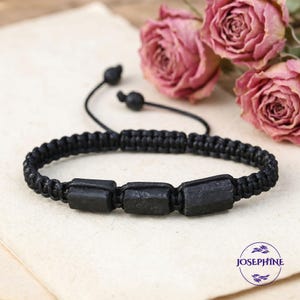 May include: Black macrame bracelet featuring three rectangular black stones. The bracelet has an adjustable sliding knot closure. The background includes dried pink roses and a cream-colored surface. The bracelet is a jewelry item.