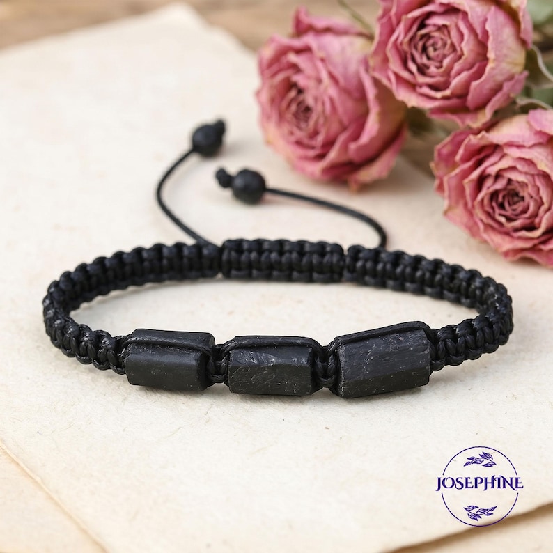 May include: Black macrame bracelet featuring three rectangular black stones. The bracelet has an adjustable sliding knot closure. The background includes dried pink roses and a cream-colored surface. The bracelet is a jewelry item.