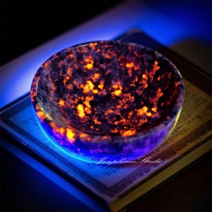 Puede incluir: Un cuenco redondo y pulido con un patrón moteado de naranja y negro. El cuenco está iluminado con un brillo azul alrededor de la base, y el interior tiene una apariencia ardiente y brillante. El texto "Josephine Studio" es visible.