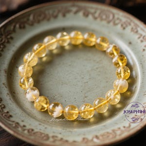 Könnte beinhalten: Ein gelbes Citrin-Armband auf einem hellgrünen Hintergrund. Das Armband besteht aus runden Perlen mit einer klaren, glasartigen Oberfläche. Die Perlen sind auf einem dünnen, goldfarbenen Draht aufgefädelt.