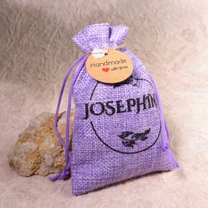 Pu&ograve; includere: Un sacchetto di juta viola con chiusura a coulisse. Il sacchetto ha un cerchio nero con la parola "JOSEPHINE" stampata in bianco. Un piccolo cartellino marrone con le parole "Handmade with love" &egrave; attaccato al sacchetto.