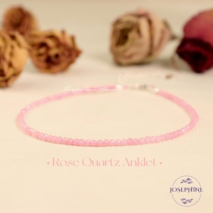 Könnte beinhalten: Ein zartes Fußkettchen aus Rosenquarz. Das Fußkettchen besteht aus kleinen, runden, hellrosa Perlen, die aneinandergereiht sind. Der Text "Rose Quartz Anklet" steht unter dem Fußkettchen. Das Josephine-Logo befindet sich unten rechts.
