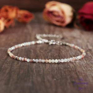 May include: A delicate beaded bracelet featuring small, faceted gemstones in shades of peach, white, and silver. The bracelet is strung on a silver chain and rests on a dark wooden surface. The bracelet is a piece of jewelry.