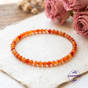 Könnte beinhalten: Ein Armband aus kleinen, runden, orangefarbenen und weißen Perlen. Das Armband wird auf einer strukturierten, cremefarbenen Oberfläche mit rosa Rosen im Hintergrund präsentiert. Es ist ein Schmuckstück.
