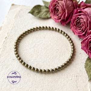 K&ouml;nnte beinhalten: Ein Armband aus goldfarbenen Perlen, bestehend aus kleinen, runden Perlen. Das Armband wird auf einer strukturierten, cremefarbenen Oberfl&auml;che mit getrockneten rosa Rosen im Hintergrund pr&auml;sentiert. Ein schlichter, eleganter Schmuck.