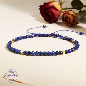 May include: A blue beaded bracelet with gold accents and a sliding knot closure. The bracelet is made of small, faceted blue beads and gold-colored square beads. The background features a red and a dried rose.