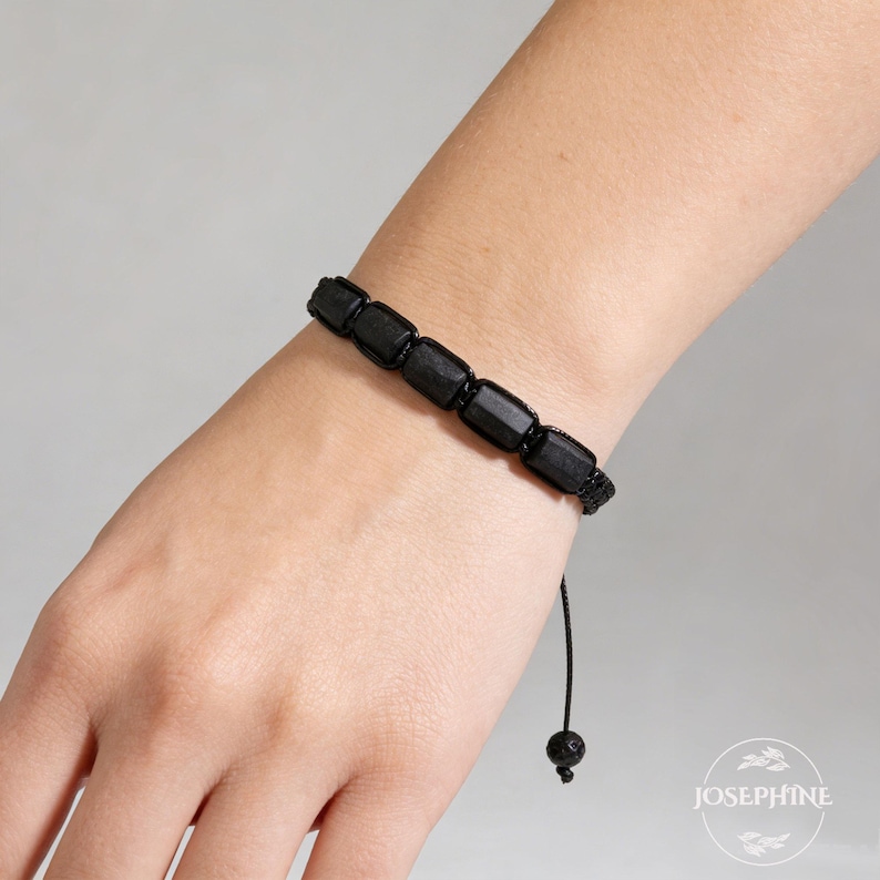 May include: Black beaded bracelet featuring rectangular beads. The bracelet is strung on a black cord with an adjustable closure. The beads appear to be made of a dark, possibly natural material. The bracelet is worn on a wrist.