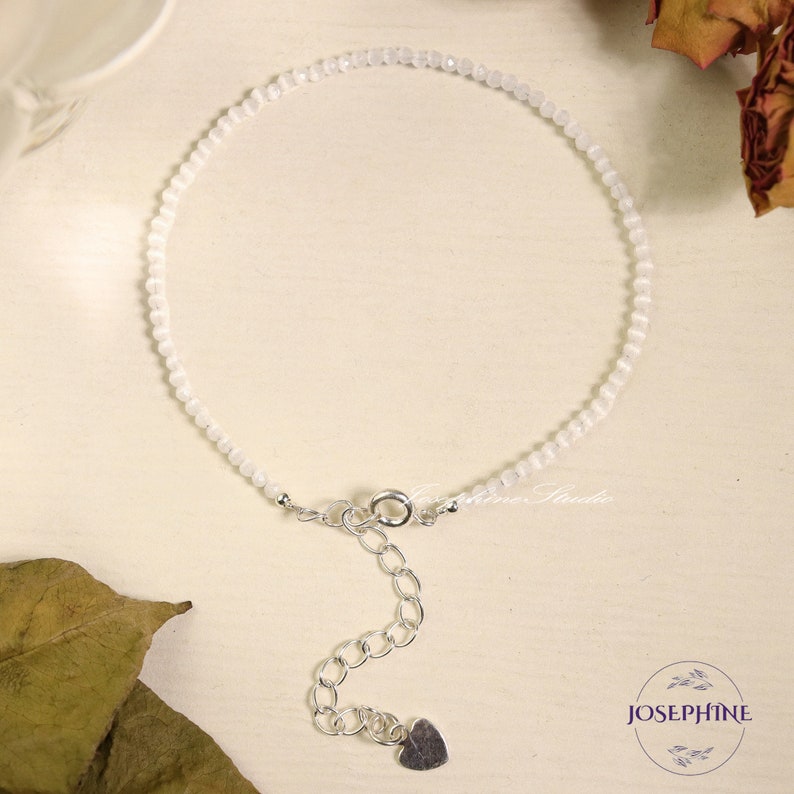 May include: A delicate white gemstone bracelet with a silver clasp and heart charm. The bracelet is made with small, round beads and has an adjustable length.