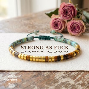 Könnte beinhalten: Ein Armband mit goldenen, braunen und türkisfarbenen Perlen, mit dem Text "STRONG AS FUCK". Das Armband liegt auf einer weißen Oberfläche, im Hintergrund sind rosa Rosen zu sehen. Unten steht der Markenname "JOSEPHINE". Ca. 18 cm lang.