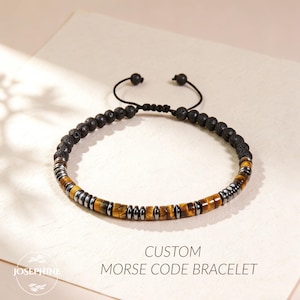Puede incluir: Pulsera ajustable de cuentas negras con cuentas de roca volcánica, cuentas de ojo de tigre y espaciadores de metal plateado. La pulsera está etiquetada como "Custom Morse Code Bracelet" con la marca "Josephine".