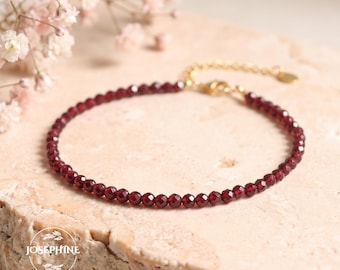 May include: A delicate bracelet featuring small, faceted, deep red garnet beads strung on a thin gold-toned chain. The piece includes a gold-toned extender chain with a small circular charm at the end. The logo Josephine is visible.