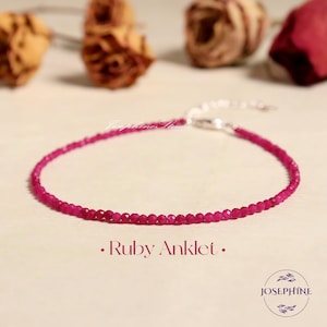 May include: A delicate ruby anklet with a silver clasp. The anklet is composed of small, faceted, deep pink gemstones. The words "Ruby Anklet" are printed below the anklet. The Josephine Studio logo is in the lower right corner.